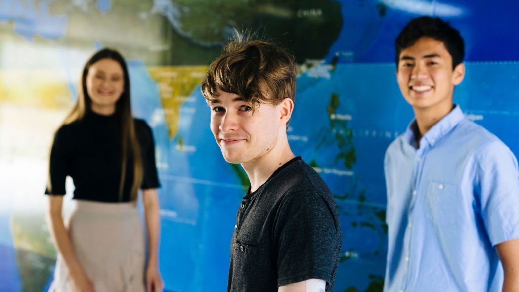 Three students stand in front of a mural of the world map, smiling at the camera. 